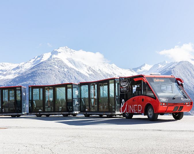 The eLiner, a modern red electric train, stands against the snow-covered backdrop of the Wildkogel Arena in Neukirchen. Majestic mountains rise up in the background.