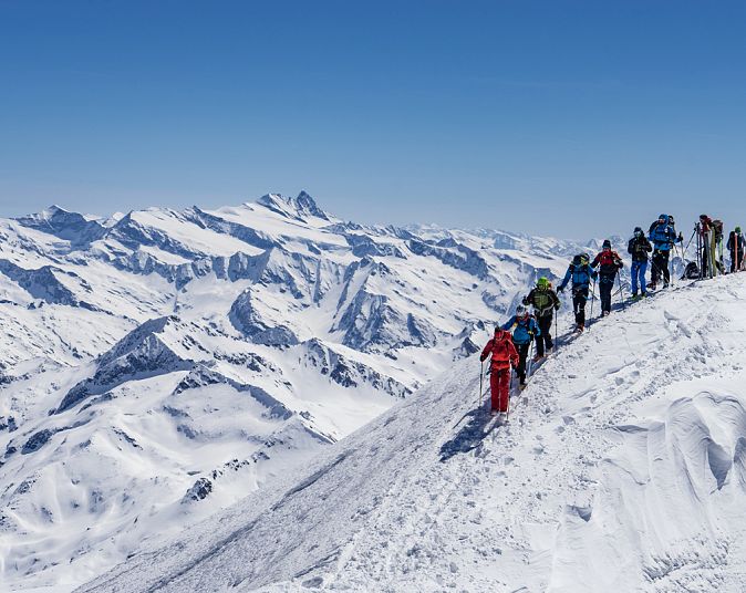 A group of people in colourful winter clothes are hiking along a snow-covered mountain ridge. In the background, majestic, snow-capped mountain peaks can be seen against a clear sky.
