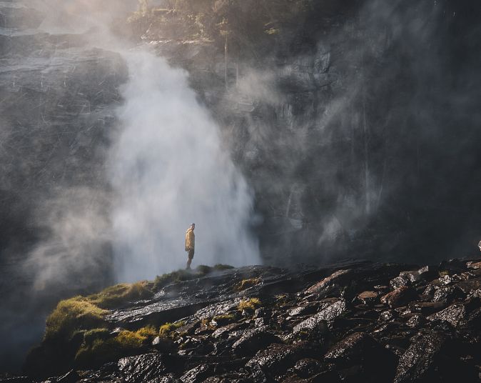 Eine Person steht auf einem felsigen Abhang, umgeben von dichtem Nebel und sich schnell bewegendem Wasserfall. Dunkle, moosbedeckte Steine schaffen eine mystische Atmosphäre.