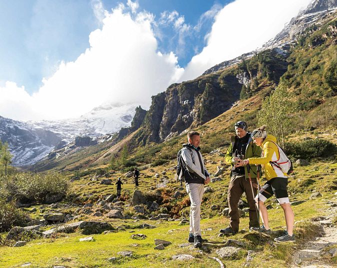 Drei Wanderer stehen auf einem grünen Pfad in den Bergen. Im Hintergrund sind schneebedeckte Gipfel und ein bewölkter Himmel zu sehen. Die Stimmung ist abenteuerlich.