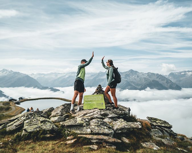 Two people are standing on a rocky mountain peak in the Wildkogel high-altitude hiking area, surrounded by clouds and mountains. They are giving each other a high-five; the sky is blue with thin clouds.