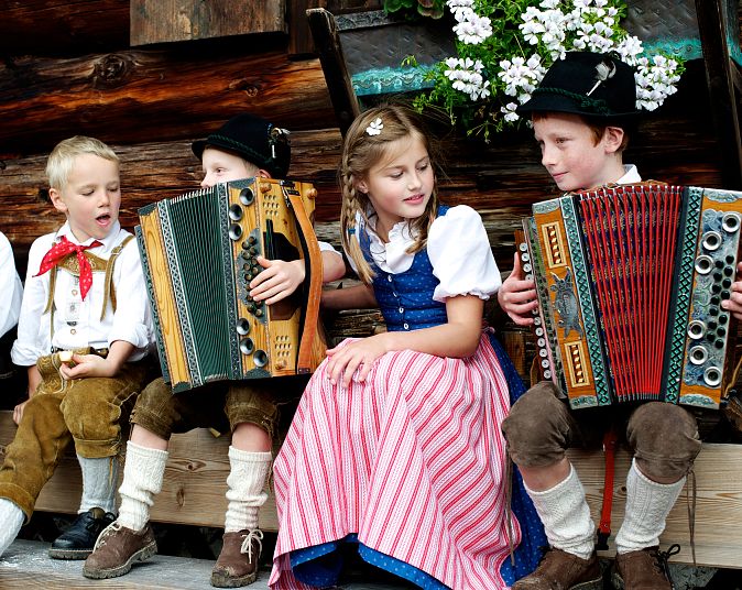 Vier Kinder in traditioneller bayerischer Kleidung sitzen auf einer Holzbank. Zwei Jungen spielen Akkordeon, während ein Mädchen und ein weiterer Junge ihnen zuhören.