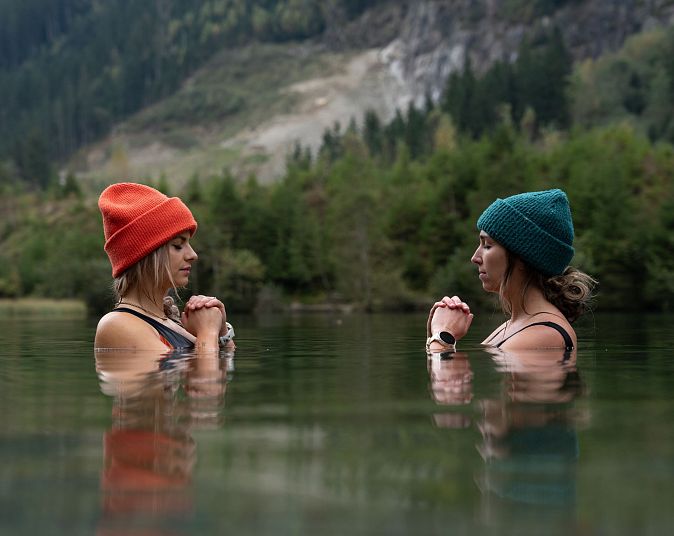 Two people wearing beanies, submerged in a calm lake surrounded by a forest, face each other with hands in prayer position. A mountainous backdrop is visible.
