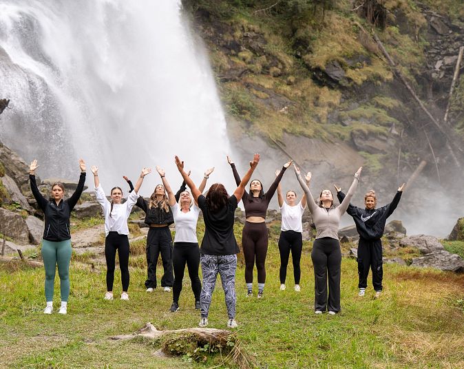 A group of people practicing yoga outdoors near a waterfall, standing on grass with their arms raised, surrounded by lush green foliage.
