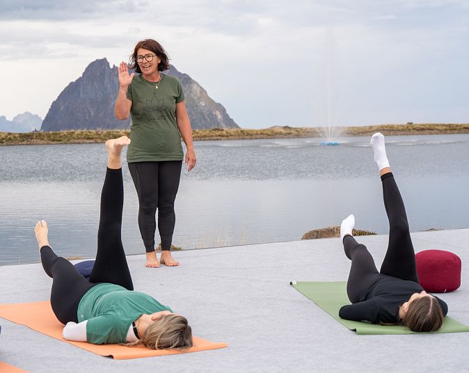 Yoga class outdoors by a lake with mountains in the background. Instructor stands while participants lie on mats, raising one leg in the air.