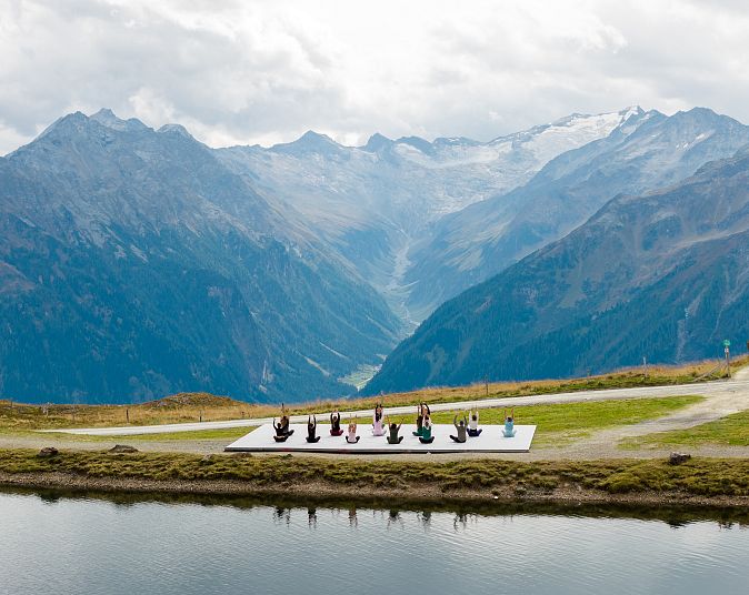 A group of people practice yoga on a platform near a serene mountain lake, surrounded by stunning alpine scenery and cloudy sky.