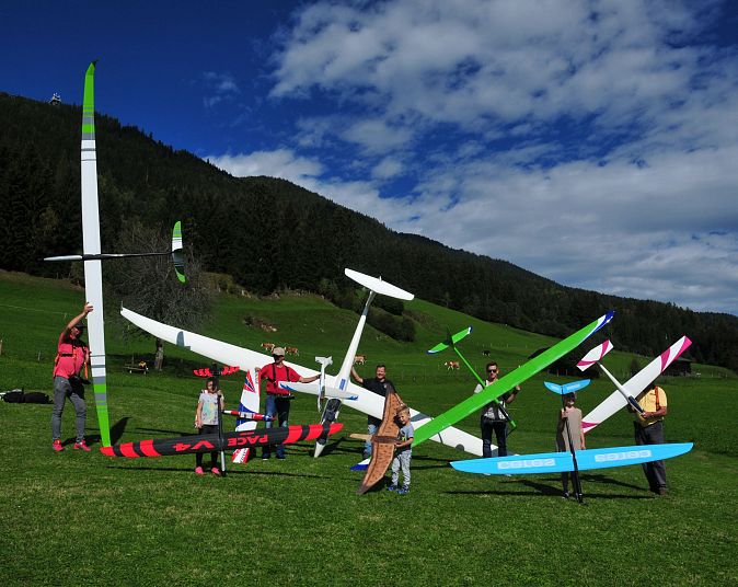 Mehrere Menschen stehen auf einer Wiese und halten bunte Modellsegelflugzeuge. Im Hintergrund sind bewaldete Hügel und ein teils bewölkter blauer Himmel zu sehen.