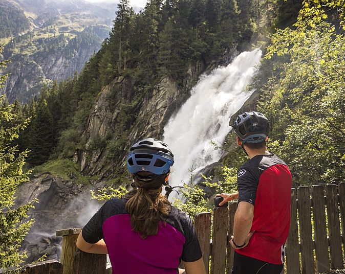 Zwei Radfahrer mit Helmen beobachten einen beeindruckenden Wasserfall, der sich durch üppige grüne Bäume und felsige Klippen schlängelt. Die Szene strahlt Abenteuerlust aus.