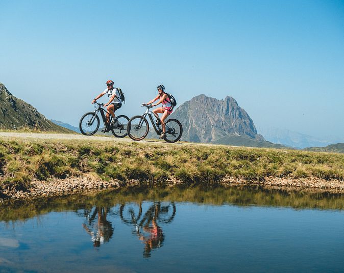 Zwei Personen fahren auf Mountainbikes entlang eines ruhigen Sees, in dem sich die umliegenden Berge spiegeln. Der Himmel ist klar und blau, und die Landschaft ist grün.