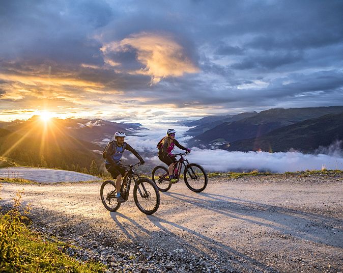 Two mountain bikers are riding along a mountain trail in the Wildkogel Arena at sunset, surrounded by dramatic skies and stunning mountain scenery.
