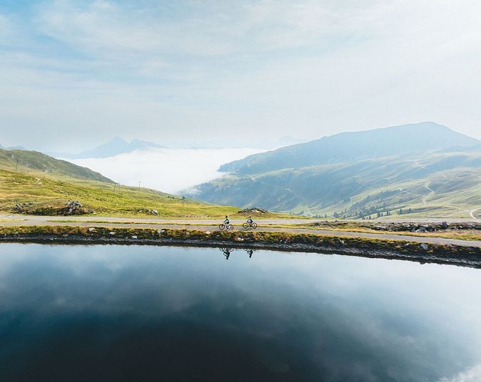 Malerische Berglandschaft in der Wildkogel-Arena mit zwei Radfahrern, die an einem ruhigen, spiegelnden Bergsee entlangfahren. Im Hintergrund beeindruckende Bergketten.