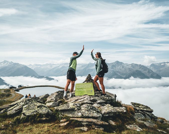 Zwei Personen machen auf einem felsigen Berggipfel einen High-Five. Umgeben von Wolken und Alpenlandschaft, unterhalten sie sich und tragen sportliche Outdoor-Kleidung.