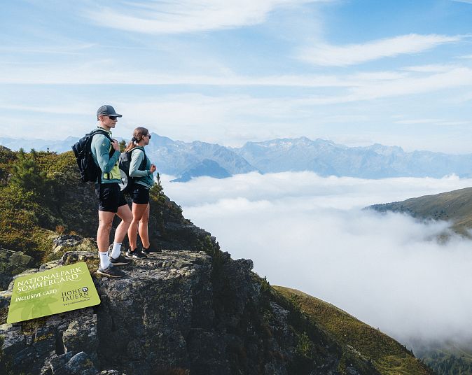 Zwei Wanderer stehen auf einem felsigen Berggipfel mit atemberaubendem Blick auf ein wolkenverhangenes Tal und ferne Bergketten unter einem klaren, blauem Himmel.
