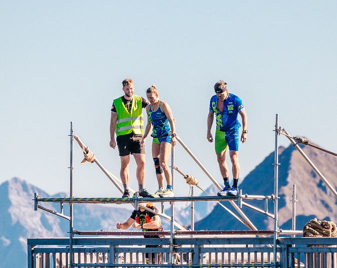 Drei Menschen stehen auf einem Baugerüst vor einer Berglandschaft, zwei in Sportkleidung, eine Person in Warnweste, darunter weitere Gerüstebene.