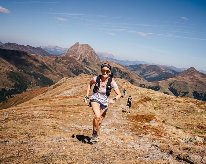 Eine Frau joggt auf einem Bergpfad mit einer beeindruckenden Berglandschaft im Hintergrund unter einem klaren blauen Himmel.