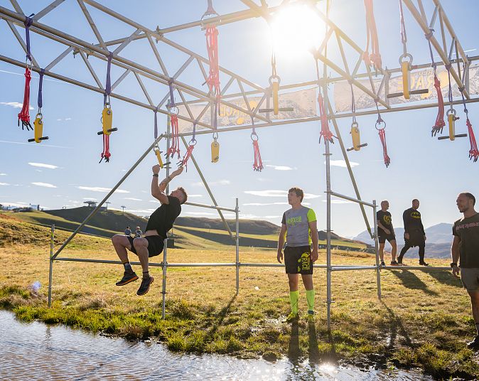 Teilnehmer eines Hindernisrennens schwingt sich an einer Metallstruktur über ein Wasserhindernis, während andere warten. Die Sonne scheint hell am Himmel.