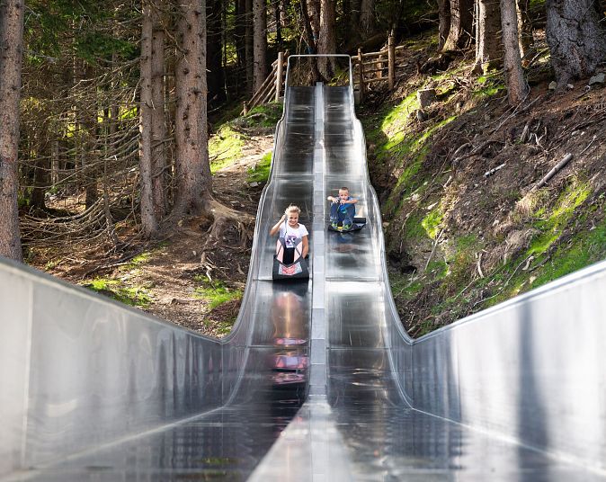 Menschen rutschen in der Wildkogel-Arena auf einer breiten Metallbahn durch einen bewaldeten Hang hinunter, umgeben von Bäumen und natürlicher Landschaft.