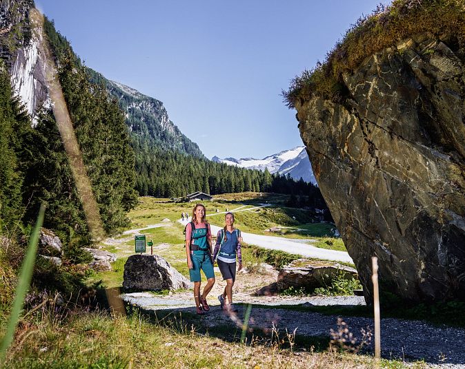 Zwei Wanderer auf einem sonnigen Pfad mit Nadelwäldern und schneebedeckten Bergen in der Wildkogel-Arena.