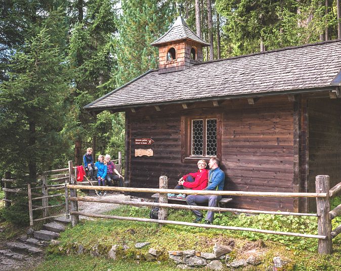 Holzhütte im Wald mit sitzenden Menschen auf einer Bank. Umgeben von hohen Bäumen und einem Holzzaun. Der Himmel ist teils sichtbar, die Atmosphäre ruhig und idyllisch.
