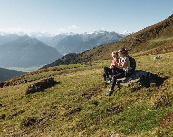 Zwei Personen sitzen auf einem Felsen in einer alpinen Landschaft. Sie genießen die Aussicht auf Berggipfel und Täler unter einem klaren blauen Himmel.