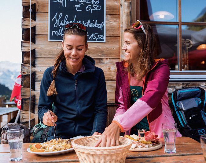 Zwei lachende Frauen essen auf einer Berghütte. Sie sitzen an einem Holztisch mit Tellern und Getränken. Im Hintergrund ist ein Bergpanorama zu sehen.