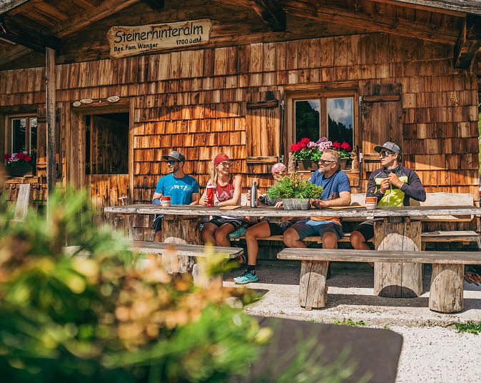 Eine Gruppe von fünf Personen sitzt an einem Holztisch vor einer Berghütte und genießt die Aussicht. Die Hütte ist aus Holz, umgeben von Grün und bepflanzt mit Blumen.