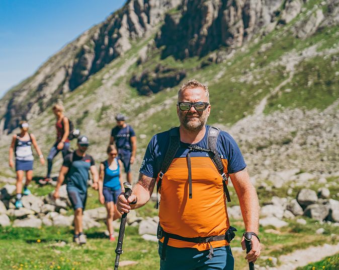 A group of hikers in colorful sportswear trekking on a rocky mountain trail with a scenic backdrop of mountains under a clear blue sky.
