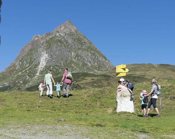 Eine Familie wandert einen grasbewachsenen Hügel hinauf. Im Hintergrund erhebt sich ein markanter Berg unter einem strahlend blauen Himmel. Wegweiser zeigen die Richtung an.