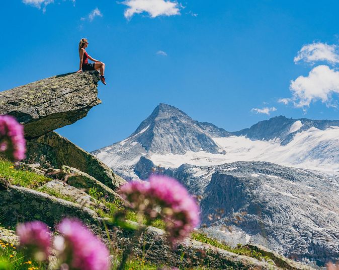 A woman is sitting on a rock overlooking snow-capped mountains under a clear sky in the Wildkogel Arena, surrounded by green countryside and flowers in the foreground.