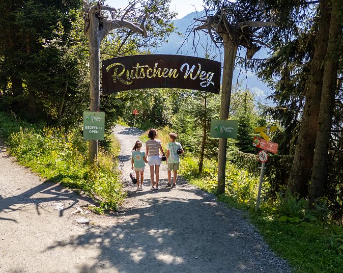 Eine Familie betritt durch einen Eingangsbogen den Rutschenweg. Der sonnige Weg ist von hohen Bäumen umgeben. Ein Schild weist auf den Beginn des abenteuerlichen Weges hin.