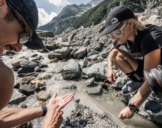 Zwei Personen erkunden felsiges Bachbett in den Bergen, sommerliche Kleidung, klare Wasserströme, grüne Hanglandschaft, helle Bergkulisse.