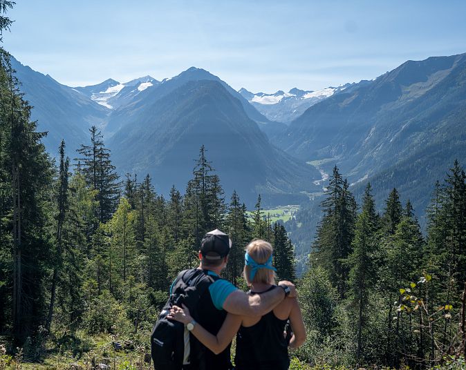 Ein Paar betrachtet eine malerische Berglandschaft mit majestätischen Gipfeln und dicht bewaldeten Hängen. Der klare Himmel verstärkt die natürliche Schönheit der Szene.