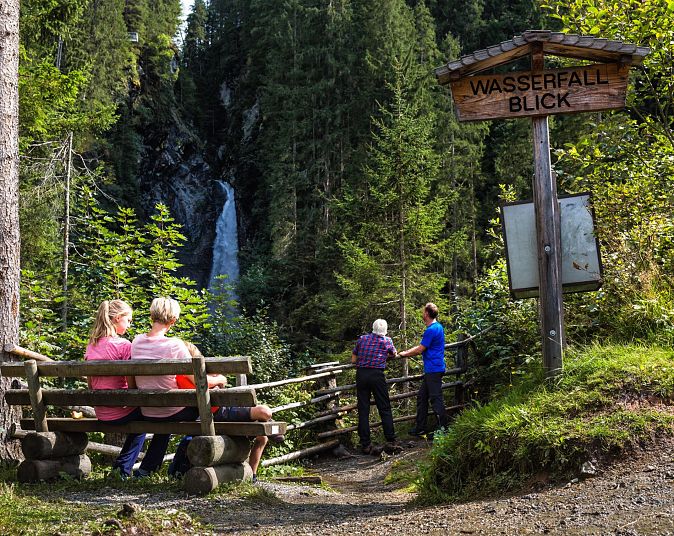 Zwei Personen sitzen auf einer Bank und betrachten den Untersulzbach-Wasserfall in einem bewaldeten Gebiet. Zwei weitere Personen stehen neben einem Schild mit der Aufschrift „Wasserfall Blick“.
