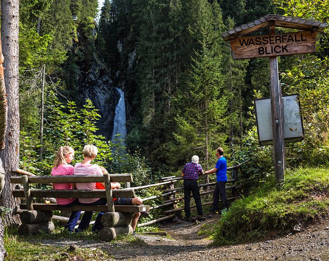 Zwei Personen sitzen auf einer Bank und betrachten den Untersulzbach-Wasserfall in einem bewaldeten Gebiet. Zwei weitere Personen stehen neben einem Schild mit der Aufschrift „Wasserfall Blick“.
