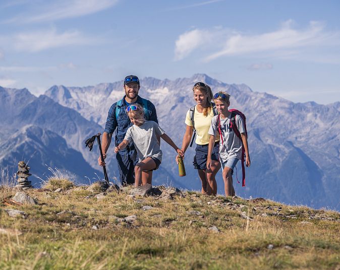 Eine Familie wandert gut gelaunt über einen sonnigen Bergrücken mit Blick auf beeindruckende, felsige Gebirge im Hintergrund, unter einem strahlend blauen Himmel.