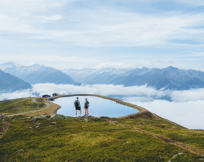 Berglandschaft mit zwei Personen, die an einem kleinen Bergsee stehen, umgeben von grüner Wiese und Bergen. Im Hintergrund sind Wolken und ein blauer Himmel zu sehen.
