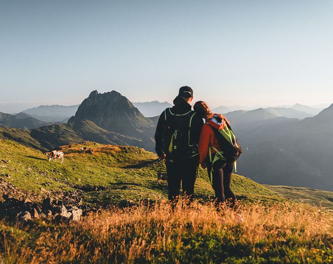 Ein Paar steht auf einem Bergpfad mit grünen Wiesen und blickt auf eine majestätische Berglandschaft. Der Himmel ist klar und die Sonne scheint sanft auf die Szenerie.