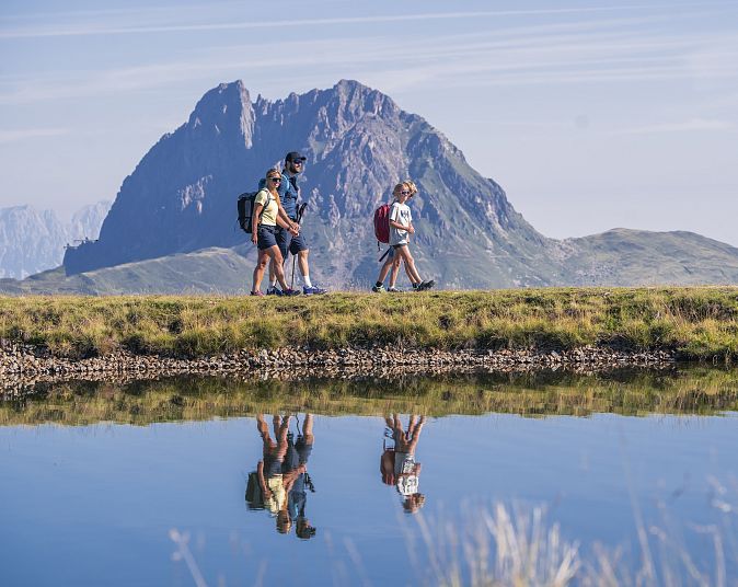 Eine Gruppe von Wanderern spaziert entlang eines Sees in den Bergen. Ihre Spiegelbilder sind im ruhigen Wasser sichtbar. Im Hintergrund erhebt sich ein markanter Gipfel.