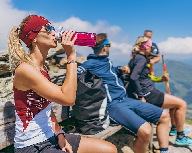 Vier Menschen entspannen auf einer Berghütte bei sonnigem Wetter. Im Vordergrund trinkt eine Frau aus einer pinken Flasche. Sie trägt Sportkleidung und eine Sonnenbrille.