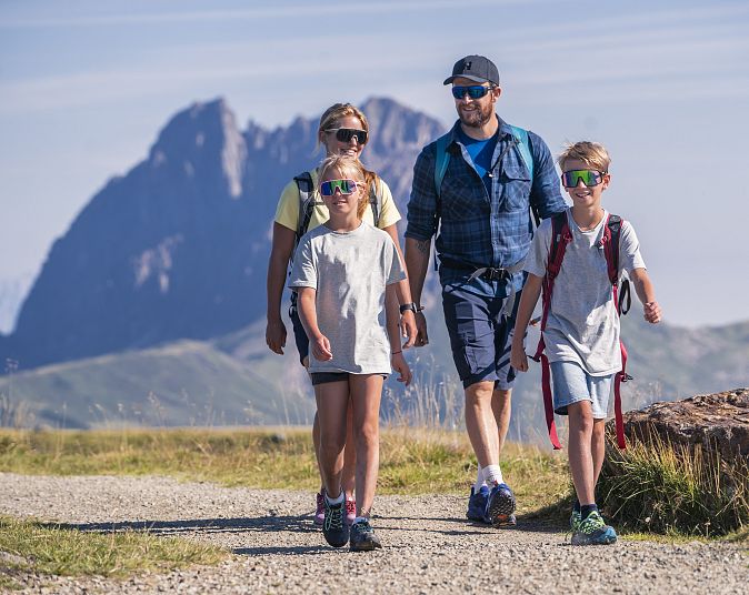 Eine Familie wandert im Höhenwandergebiet Wildkogel an einem sonnigen Tag. Im Hintergrund ist der markante Große Rettenstein zu sehen. Sie tragen Freizeitkleidung und Rucksäcke.