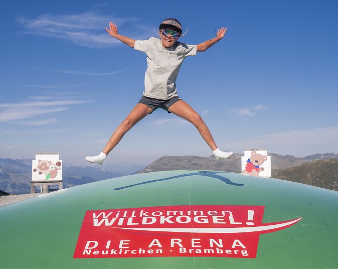Ein Kind springt mit ausgestreckten Armen und Beinen auf dem Riesentrampolin in der Abenteuer-Arena Kogel-Mogel. Im Hintergrund sind Berge und blauer Himmel zu sehen. Ein Schild sagt "Willkommen Wildkogel".