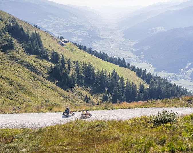 Zwei Personen fahren mit Mountaincarts die  kurvige Mountaincart-Strecke in der Wildkogel-Arena hinunter, umgeben von grünen Hügeln und einem weiten, nebeligen Tal im Hintergrund.