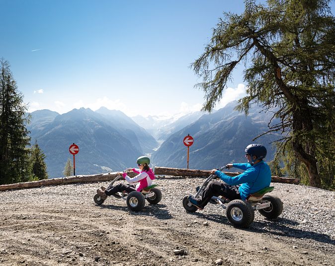 Zwei Personen fahren bei sonnigem Wetter auf geländetauglichen Mountaincarts einen Schotterweg entlang, umgeben von Bergen und Bäumen, mit einem weiten Tal im Hintergrund.