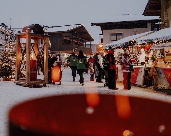 Verschneiter Weihnachtsmarkt mit festlich dekorierten Ständen, leuchtenden Lichtern und Menschen in Winterkleidung. Im Vordergrund unscharfe Lichter, im Hintergrund Holzhäuser.