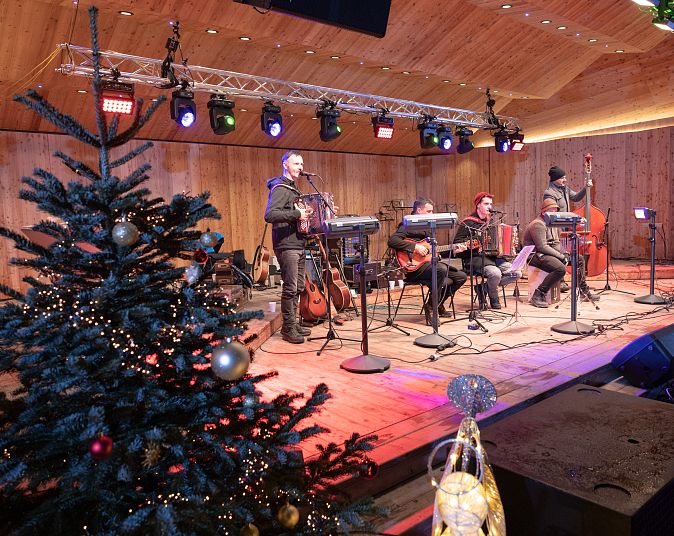 Eine Band spielt auf einer Holzbühne unter bunten Lichtern. Ein geschmückter Weihnachtsbaum steht im Vordergrund, der Raum wirkt festlich und einladend.