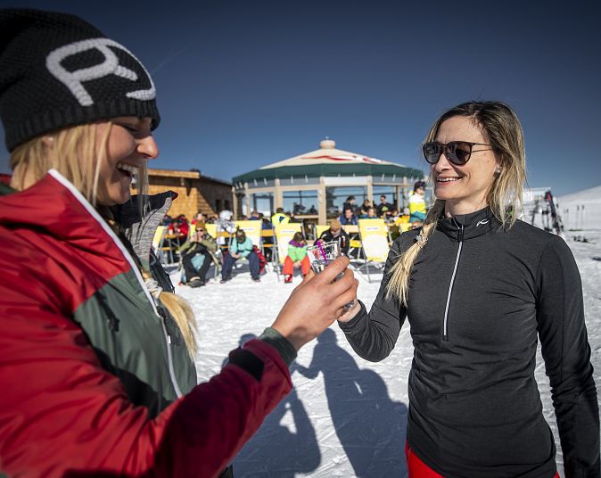 Zwei Frauen in Winterkleidung lächeln sich an und stoßen mit Getränken an. Im Hintergrund sind eine Skihütte und eine Gruppe von Leuten auf Liegestühlen im Schnee zu sehen.