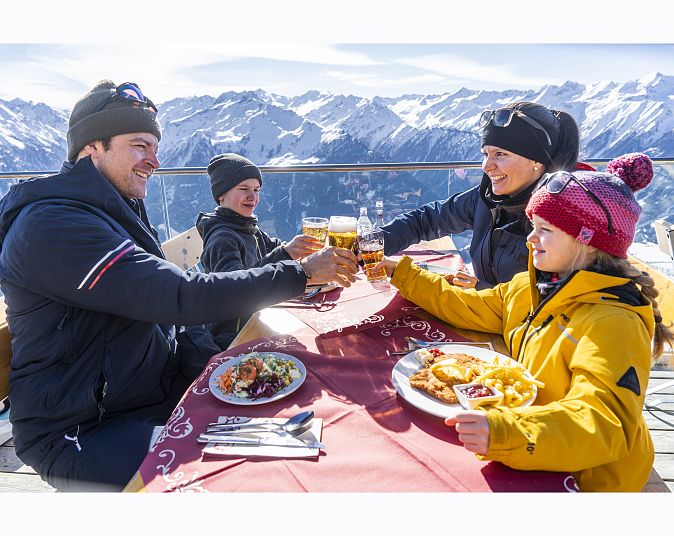 Eine Familie sitzt an einem Tisch auf einer winterlichen Terrasse, umgeben von schneebedeckten Bergen. Sie genießen gemeinsam Essen und stoßen mit Getränken an.