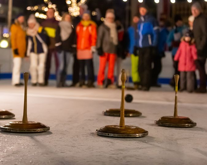 Personen spielen Eisstockschießen auf einer Eisbahn, während eine Gruppe von Zuschauern warm angezogen zuschaut. Der Fokus liegt auf den Eisstöcken im Vordergrund.