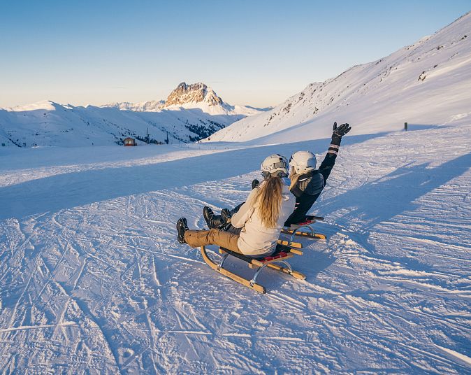 Zwei Personen rodeln auf einem schneebedeckten Hang in den Alpen. Sie tragen Winterkleidung und Helme. Im Hintergrund sind Berggipfel und ein strahlend blauer Himmel zu sehen.