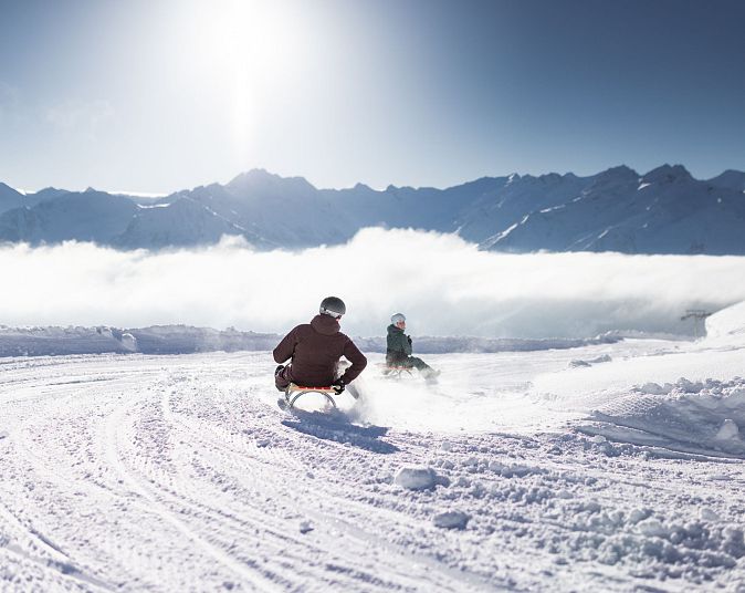 Zwei Personen rodeln bei Sonnenschein auf einer verschneiten Piste in den Bergen. Sie sind auf der längsten beleuchteten Rodelbahn der Welt in der Wildkogel-Arena.